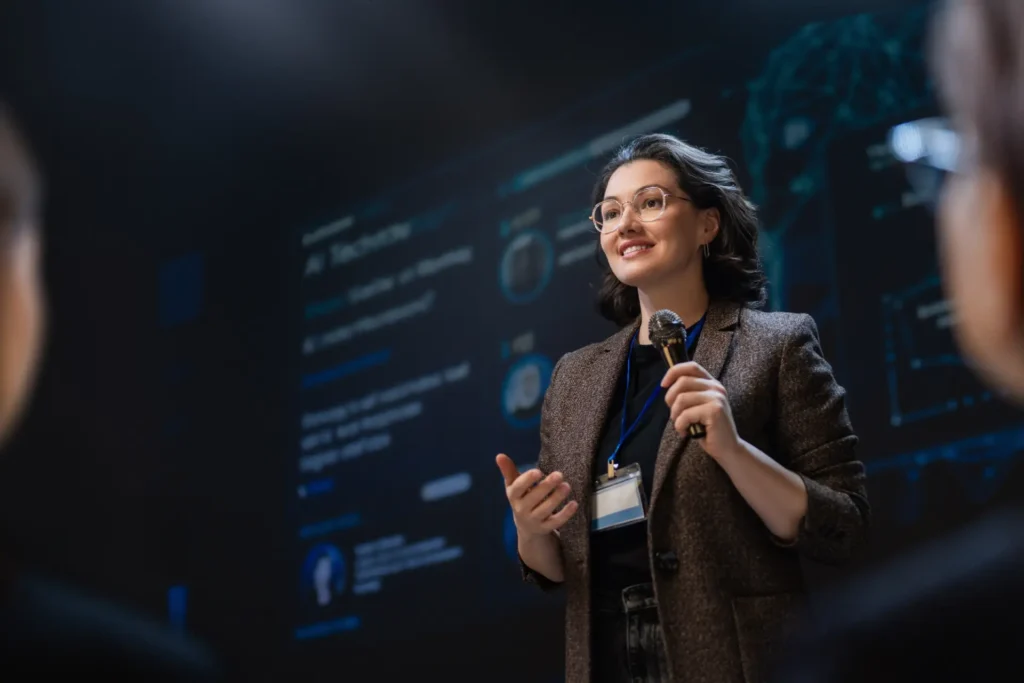 Professional speaker presenting on stage with microphone and digital interface background — CXbuildr office design and construction services.